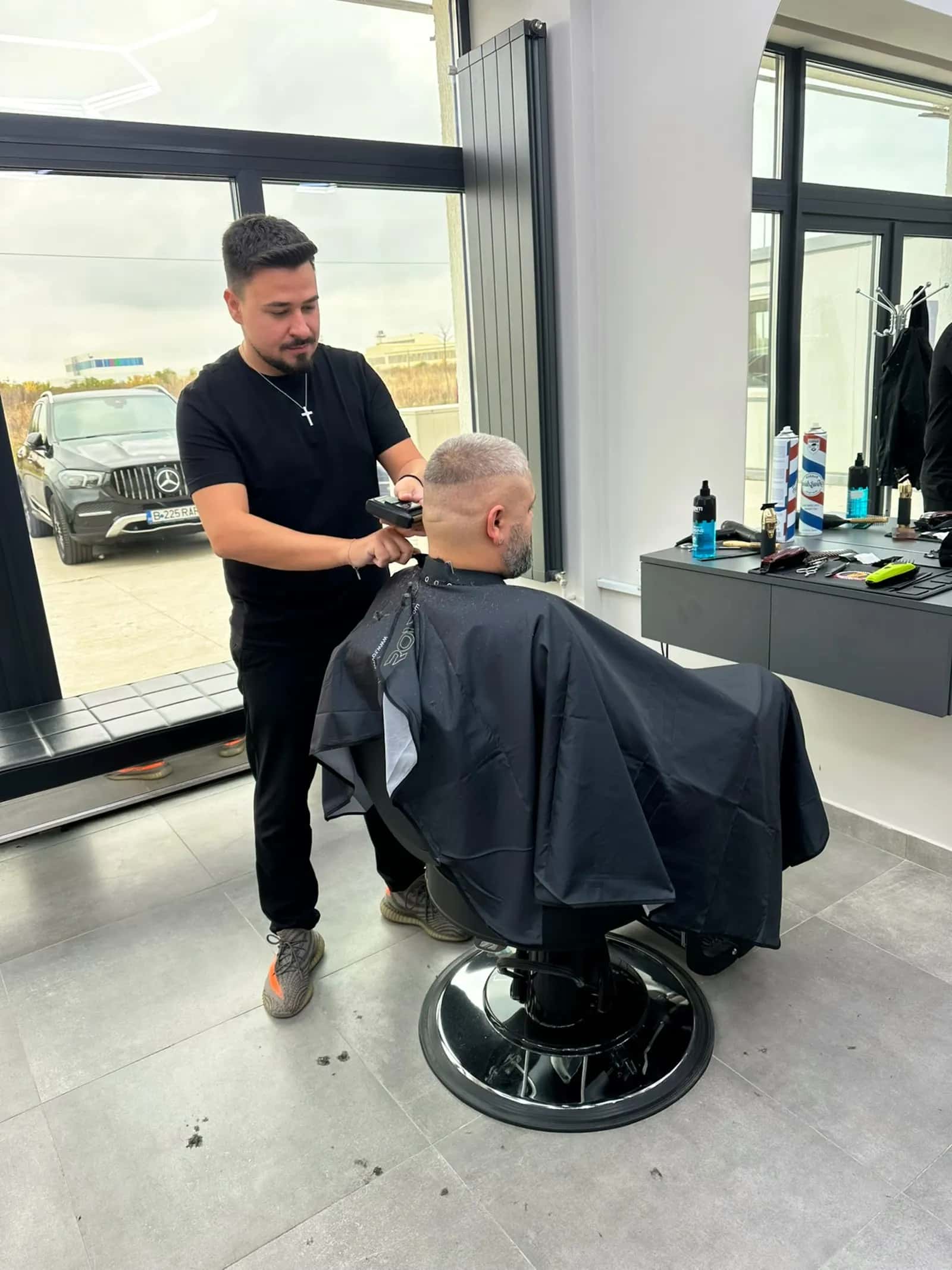 Frizer lucrând un skin fade cu mașina, lângă geamul salonului