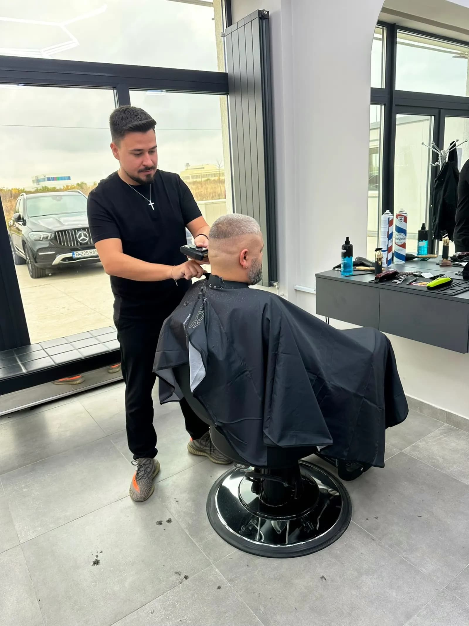 Frizer lucrând un skin fade cu mașina, lângă geamul salonului